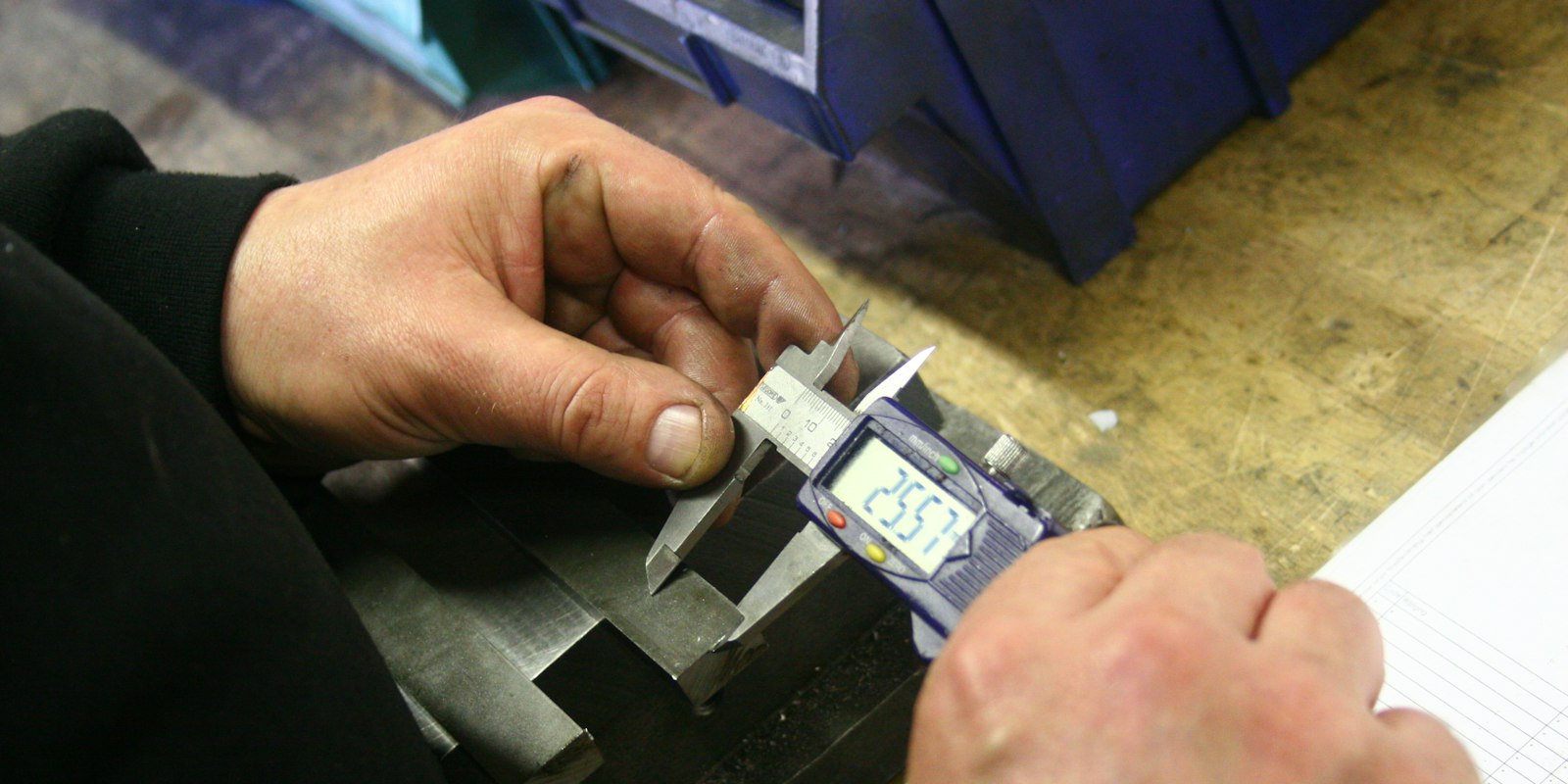 Hands using a digital caliper to measure a machined metal part on a workbench.