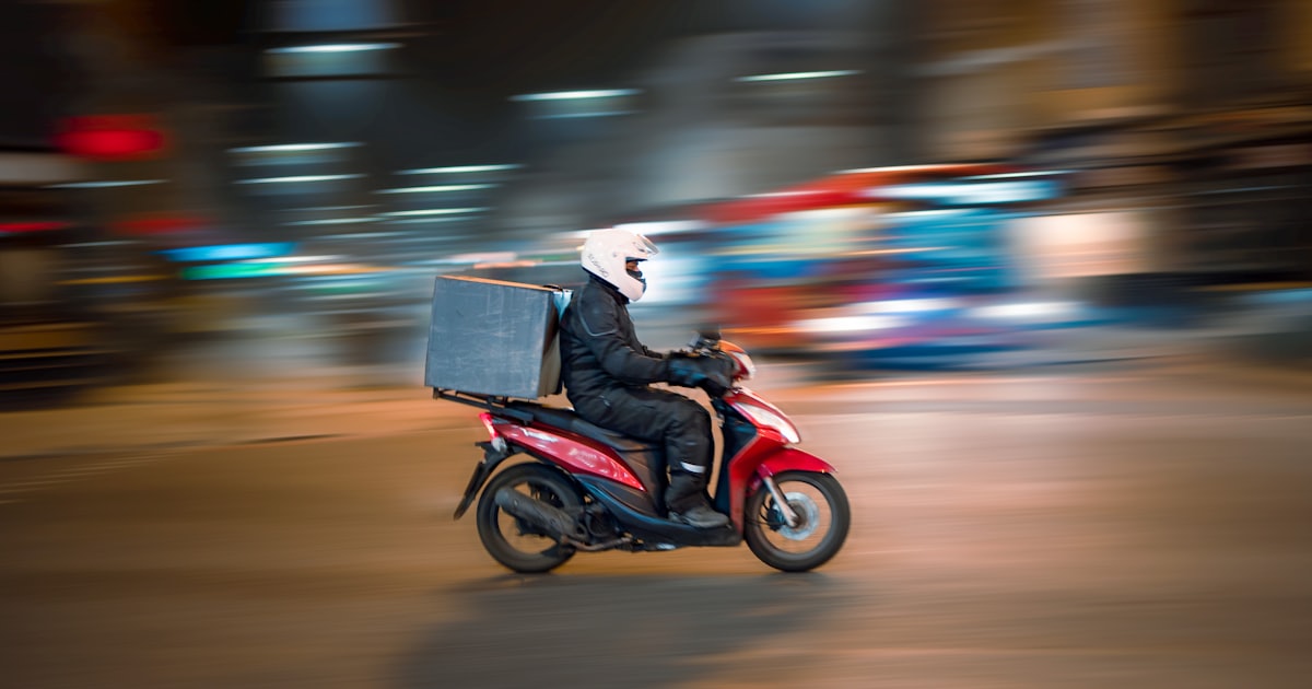 A delivery driver on a red scooter riding through a city at night