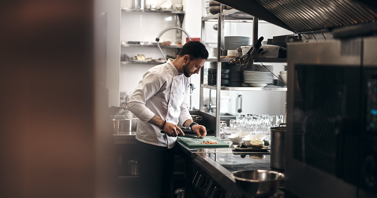 A chef in a white coat chopping vegetables on a green board in a kitchen