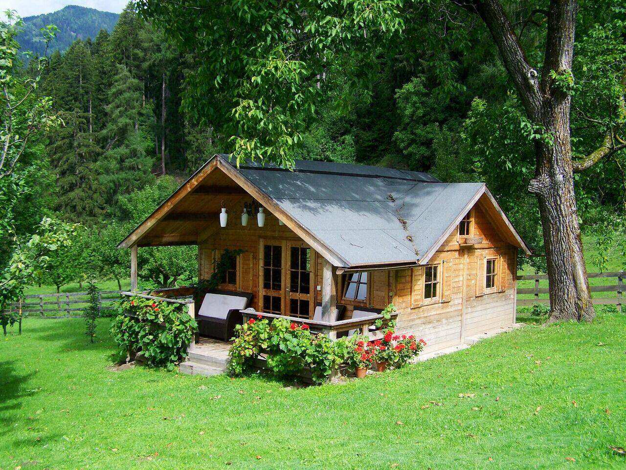 A small timber-clad garden suite with a covered porch and gable roof, set among trees at the back of a residential lot