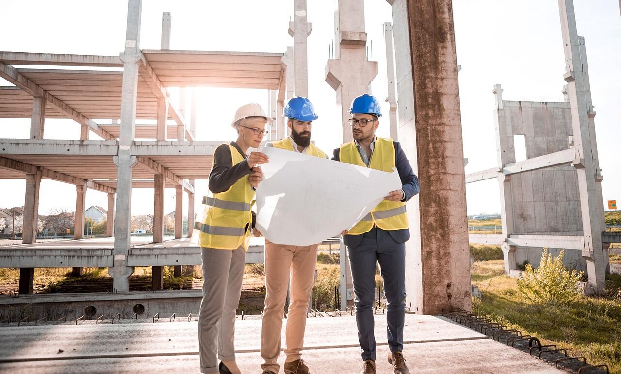 Three people in hard hats and hi-vis vests reviewing a full-size drawing at a concrete-frame construction site