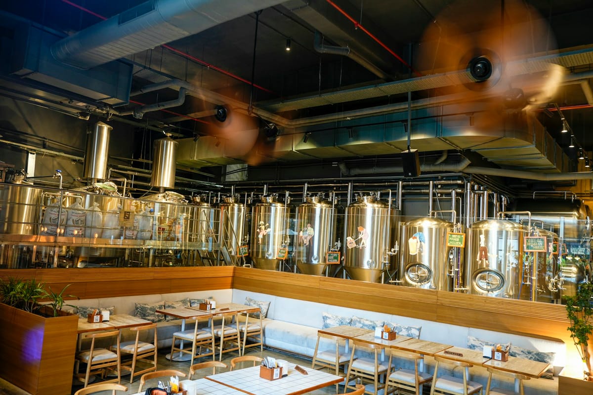Wide shot of the taproom: communal timber tables and the bar with the gleaming brew tanks in the background