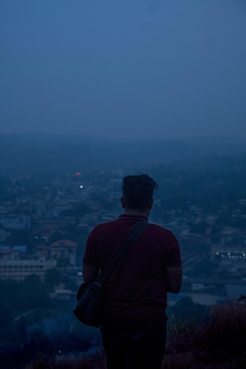 A figure seen from behind on a hillside above a city at blue hour, face not visible