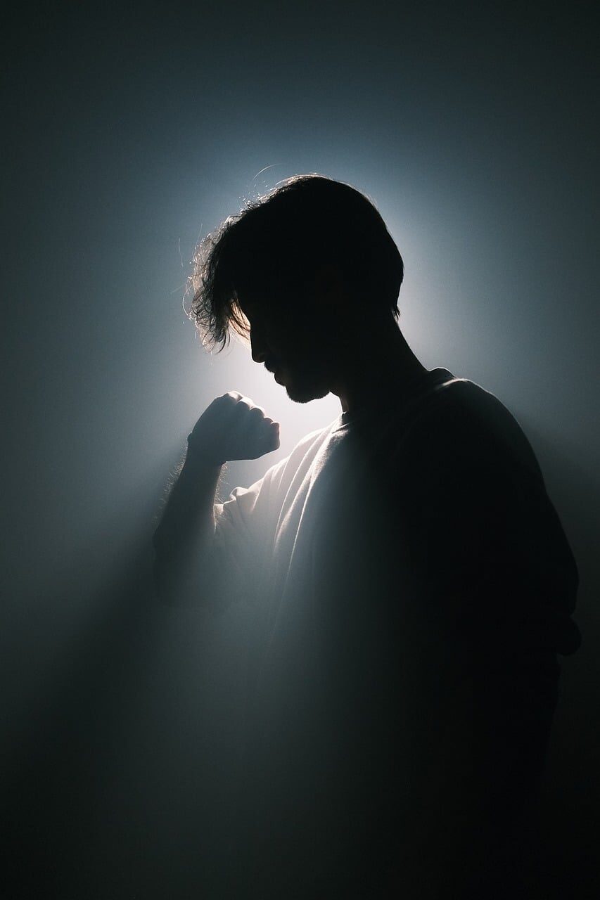 A silhouetted figure with a hand raised to the face, rimmed by a single hard backlight through haze