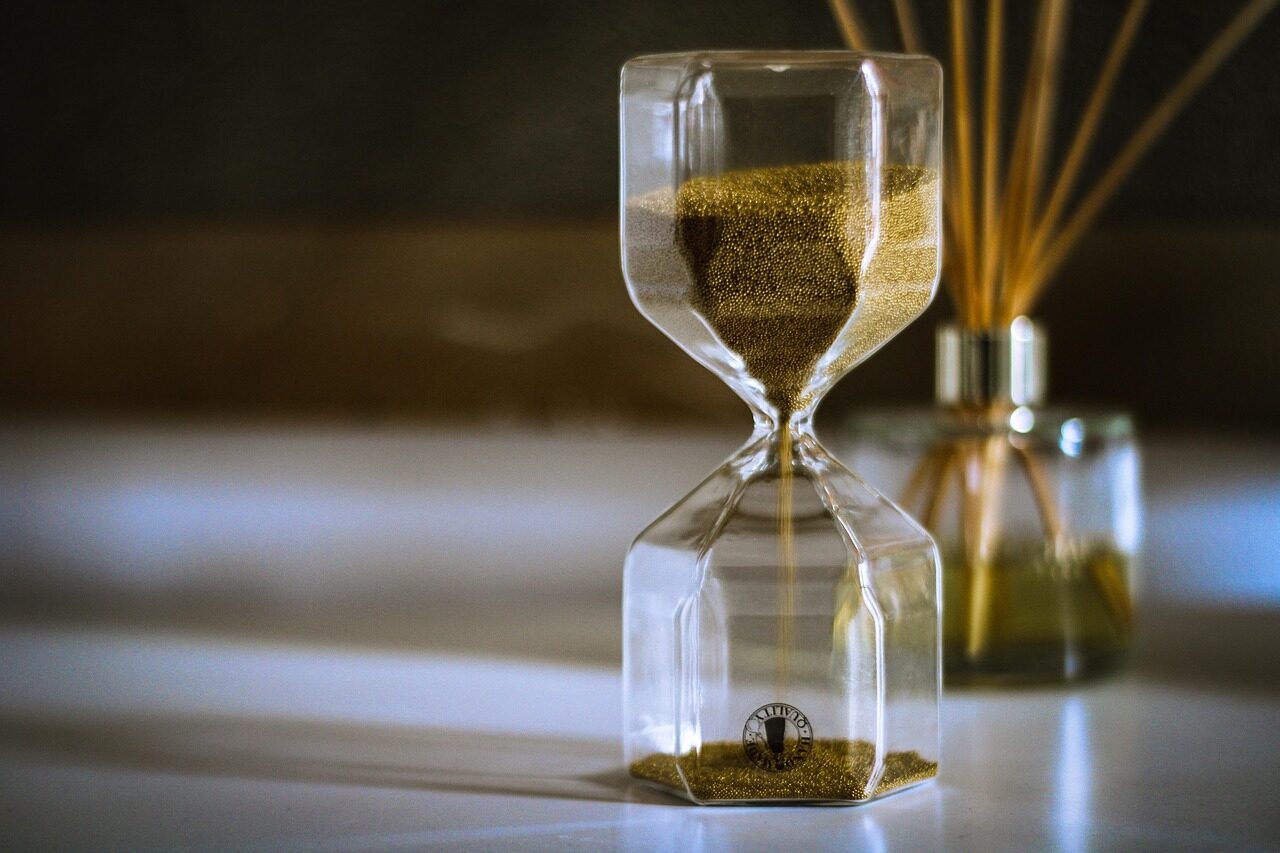 An hourglass on a table in warm backlight, the sand mid-fall and the background thrown out of focus