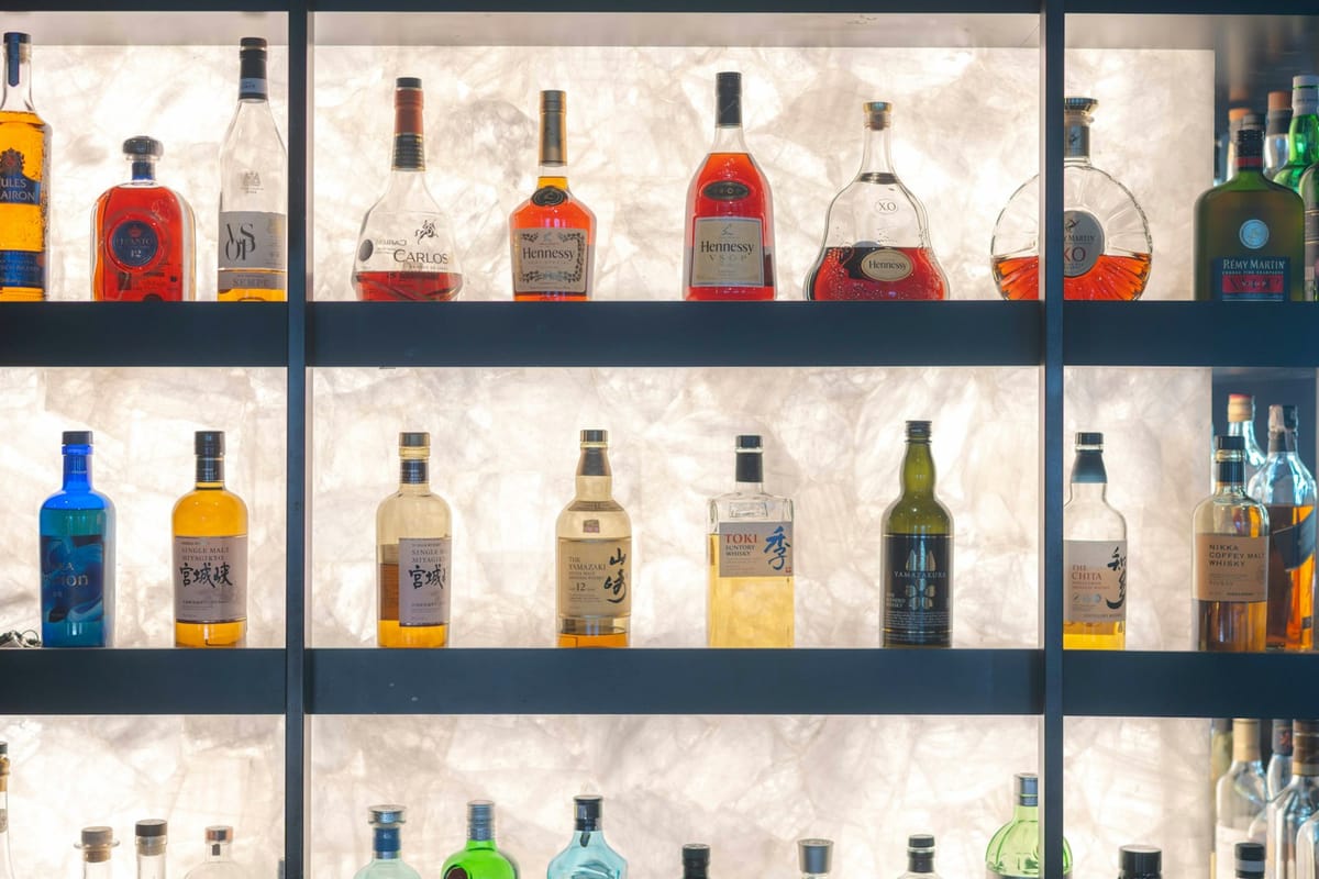 The Velvet Cipher bar counter at night — rows of rare spirits backlit against noir-black walls