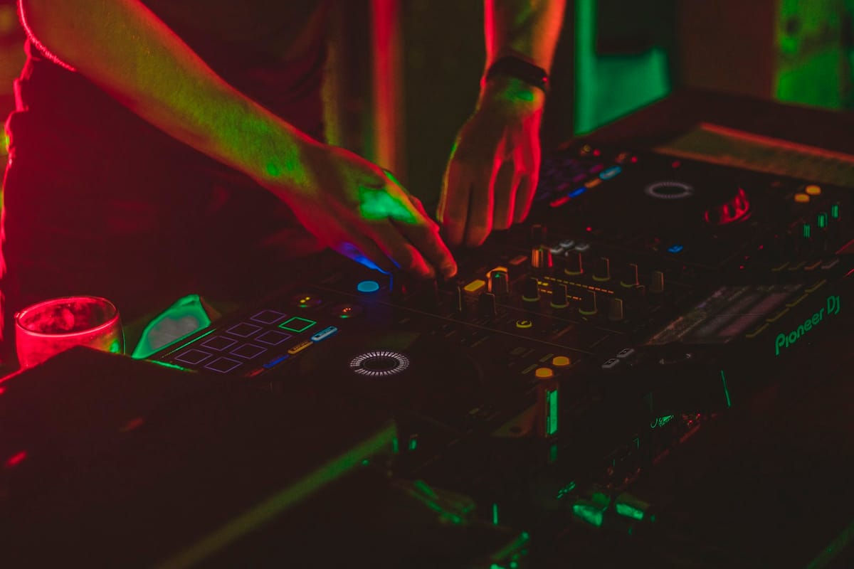 The DJ at the booth — silhouette backlit by coloured light, mixing in the dark room