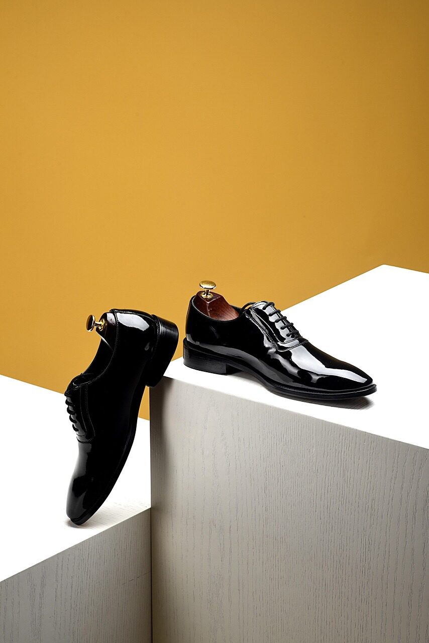 A pair of black patent oxford shoes arranged on a white plinth against an ochre background
