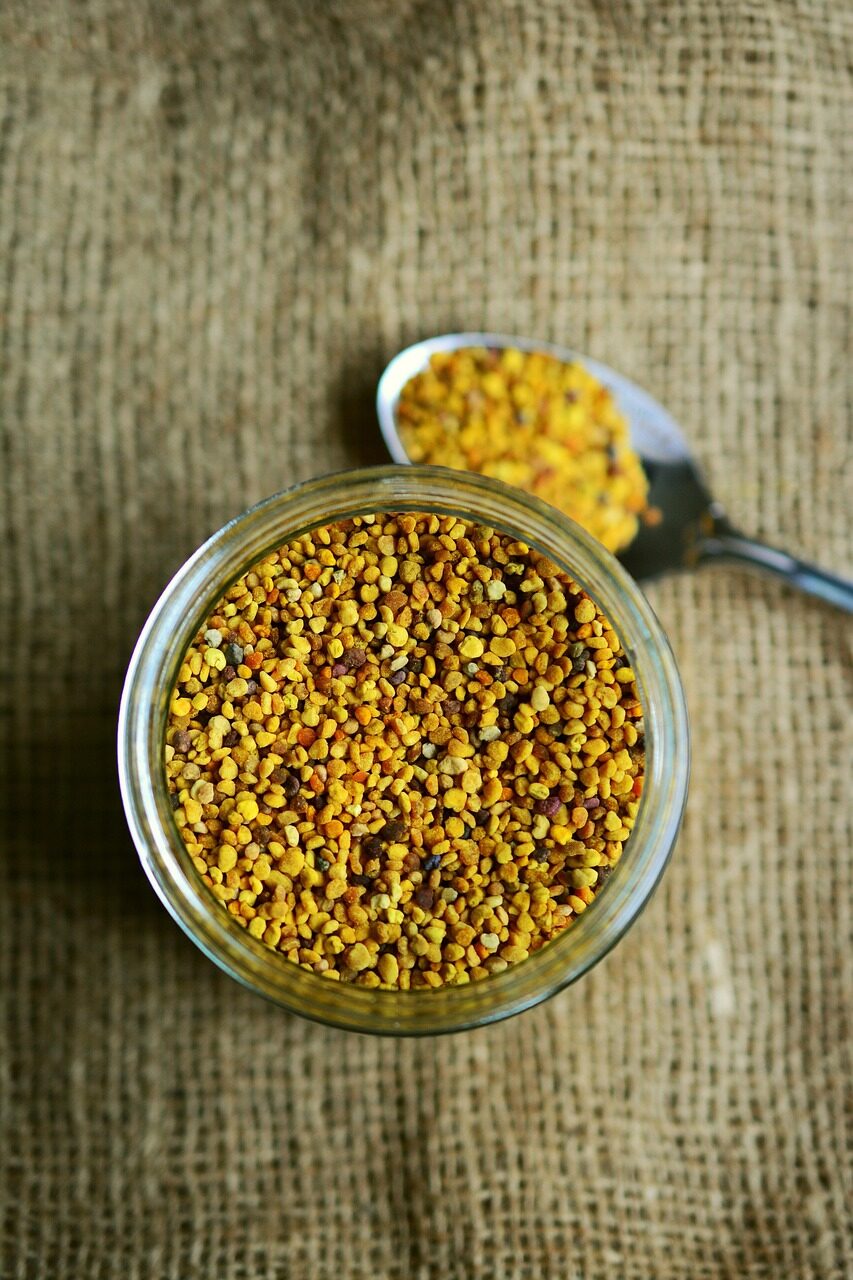 Bee pollen granules in a glass bowl beside a spoonful, on woven hessian cloth
