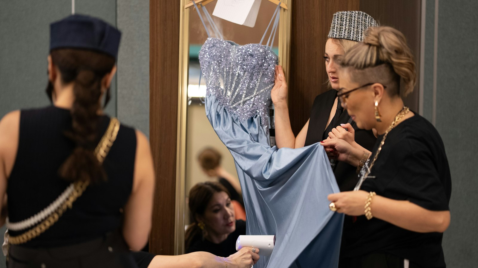 Two dressers adjusting a beaded blue gown on a hanger backstage, with a performer waiting beside them