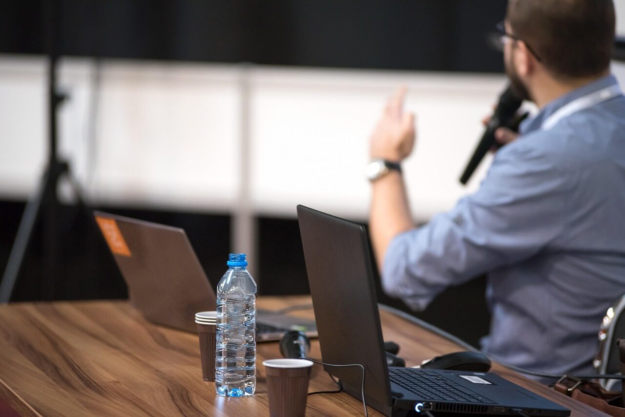 A panellist holding a handheld microphone at a table of open laptops during a session