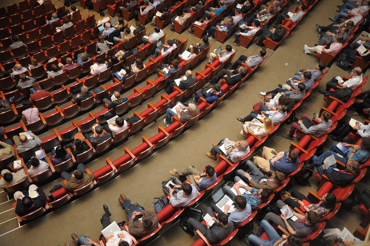 A packed auditorium of red seats photographed from a high side balcony
