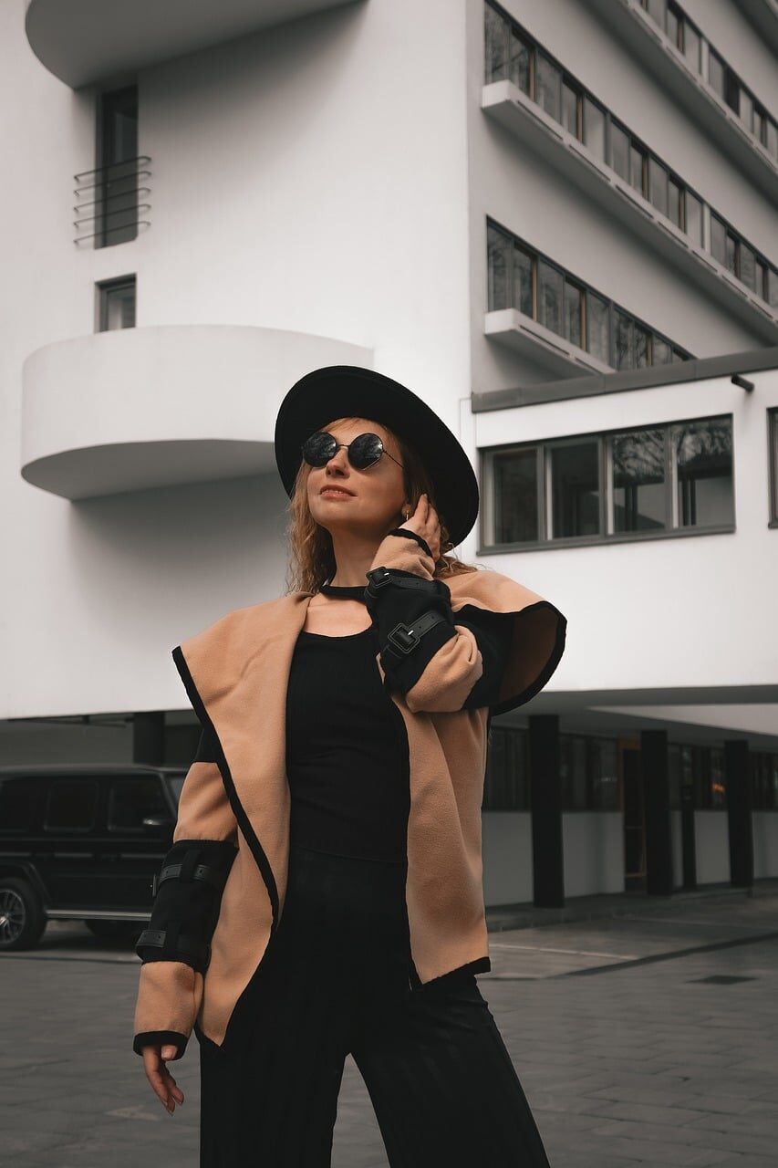A woman in a camel coat, black wide-brimmed hat and sunglasses standing before a white modernist building