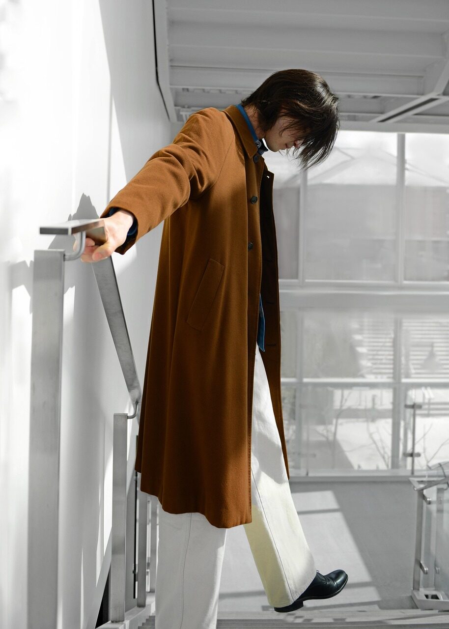 A figure in a long tan coat on a white balcony, one arm outstretched along the rail