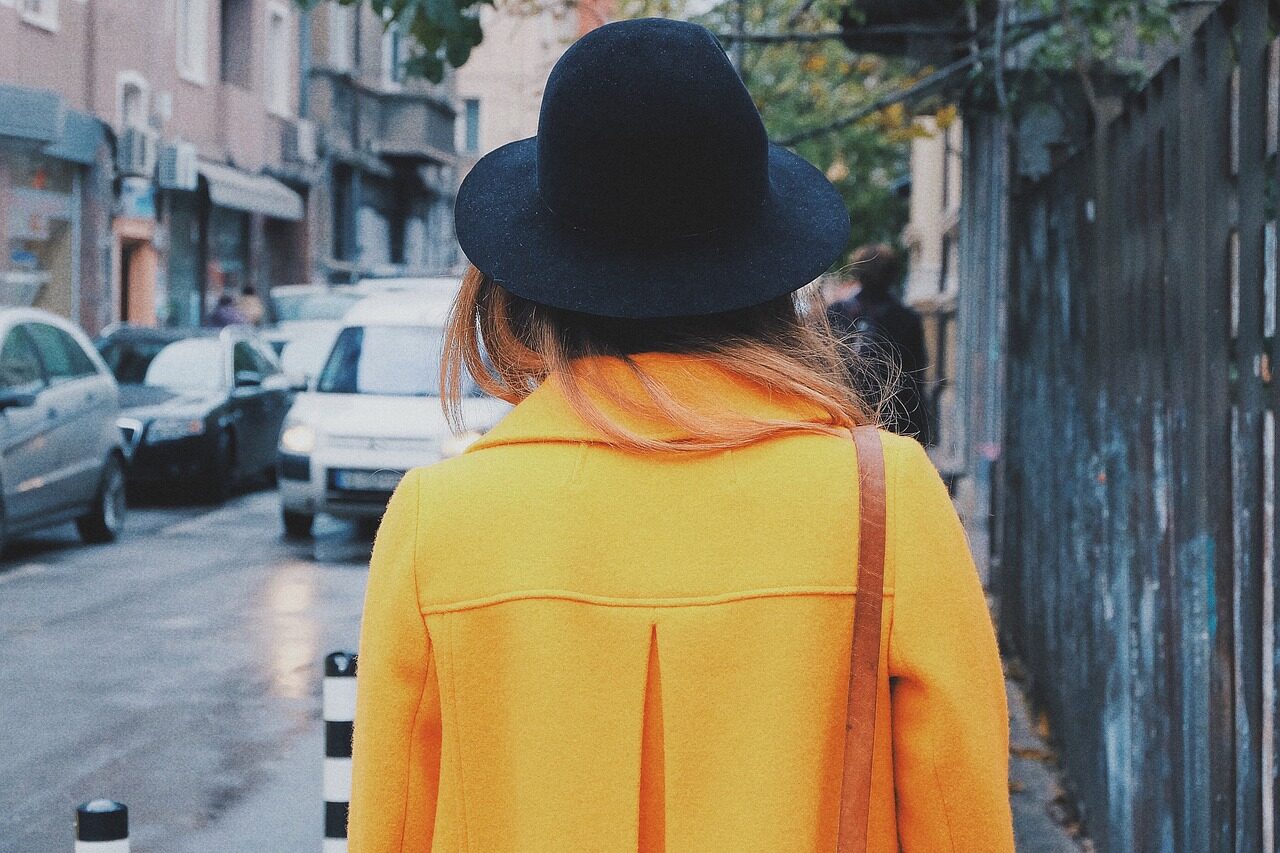 A woman photographed from behind in a yellow coat and navy wide-brimmed hat on a city street