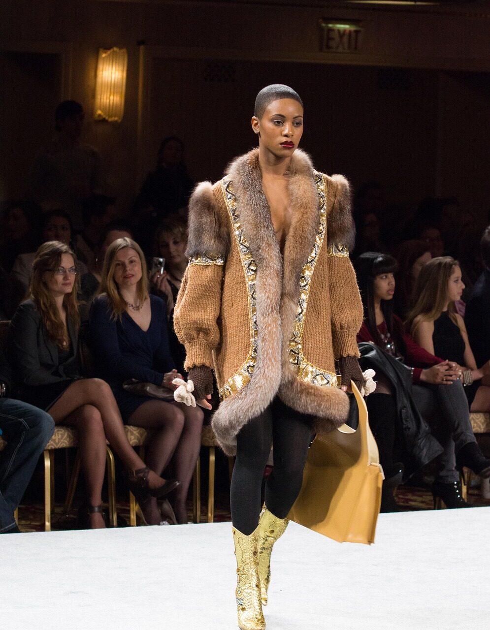 A model in a shearling-trimmed coat and gold boots walking a catwalk past a seated audience