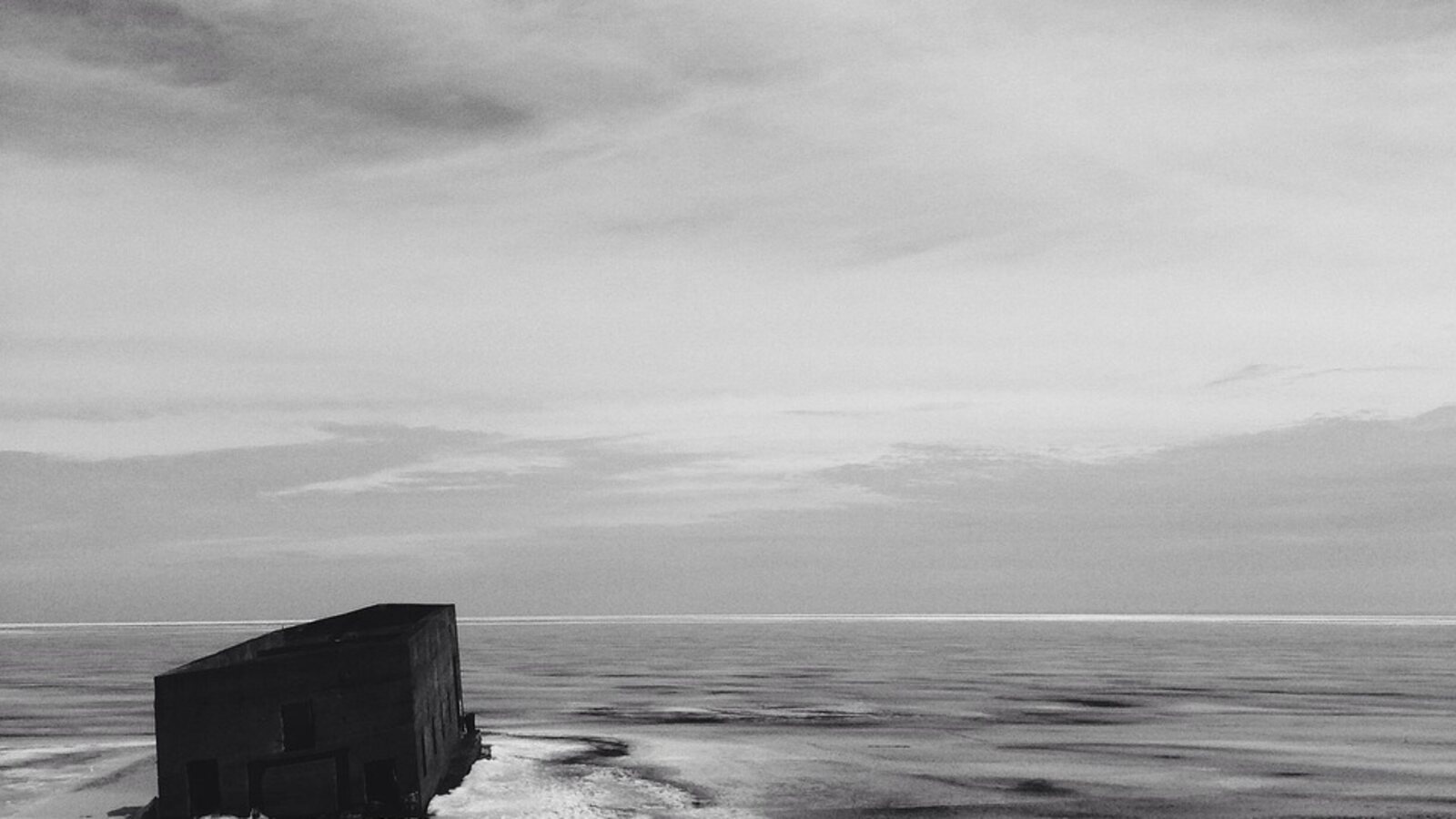 A frozen lake in black and white: a dark timber crib sitting out on the ice near the shore, under a wide flat horizon and streaked cloud
