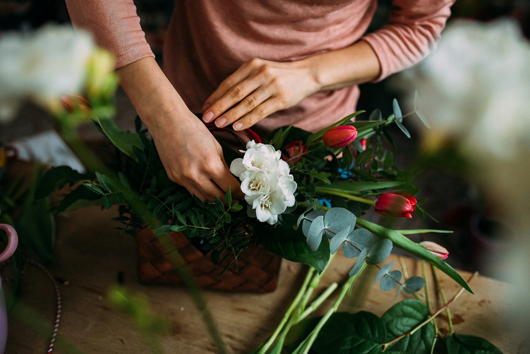A florist's hands arranging a loose bouquet of white blooms, red tulips and eucalyptus stems at a workbench