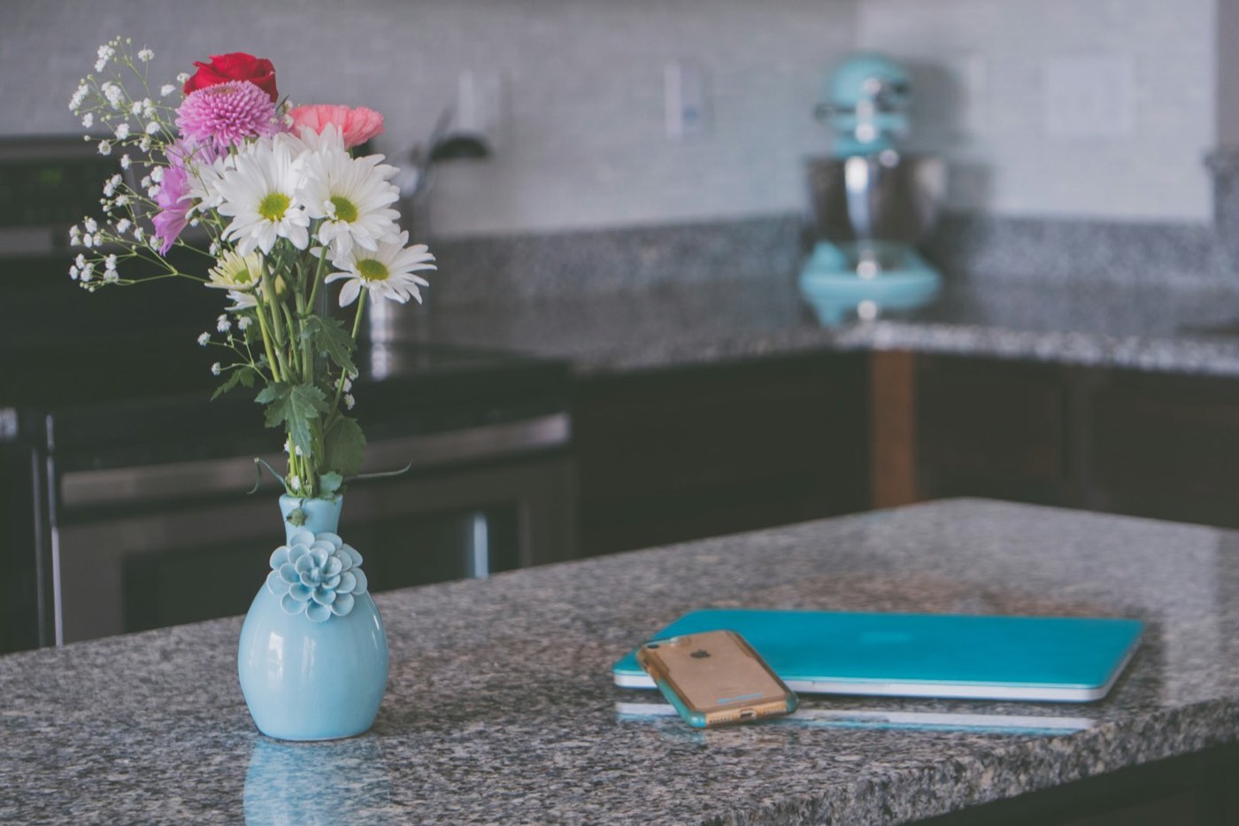 A small pale blue ceramic vase holding daisies, a red rose and pink blooms on a kitchen counter