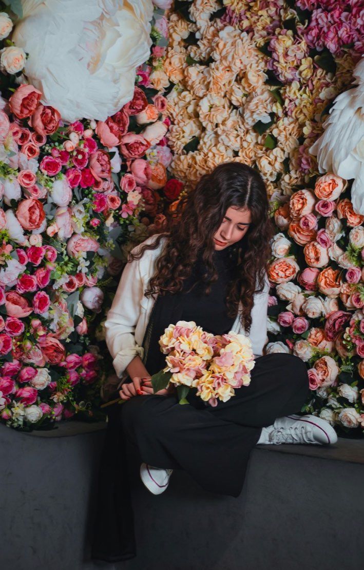Portrait of a woman seated amid a wall of flowers, holding a small bouquet, representing Teresa Nakamura