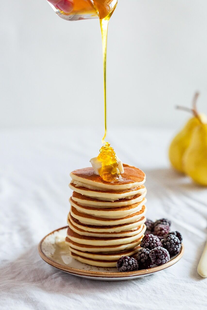 A tall stack of pancakes with honey being poured over the top against a white background