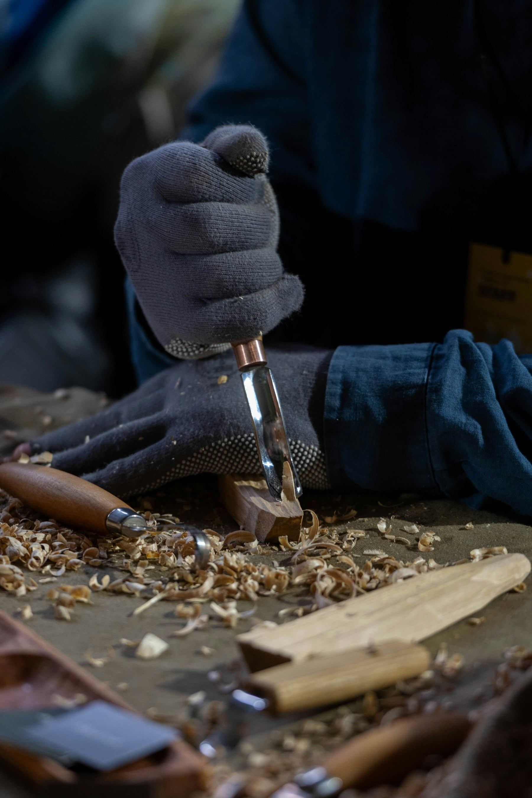 A gloved hand guiding a chisel through a wood joint, shavings curling off the blade