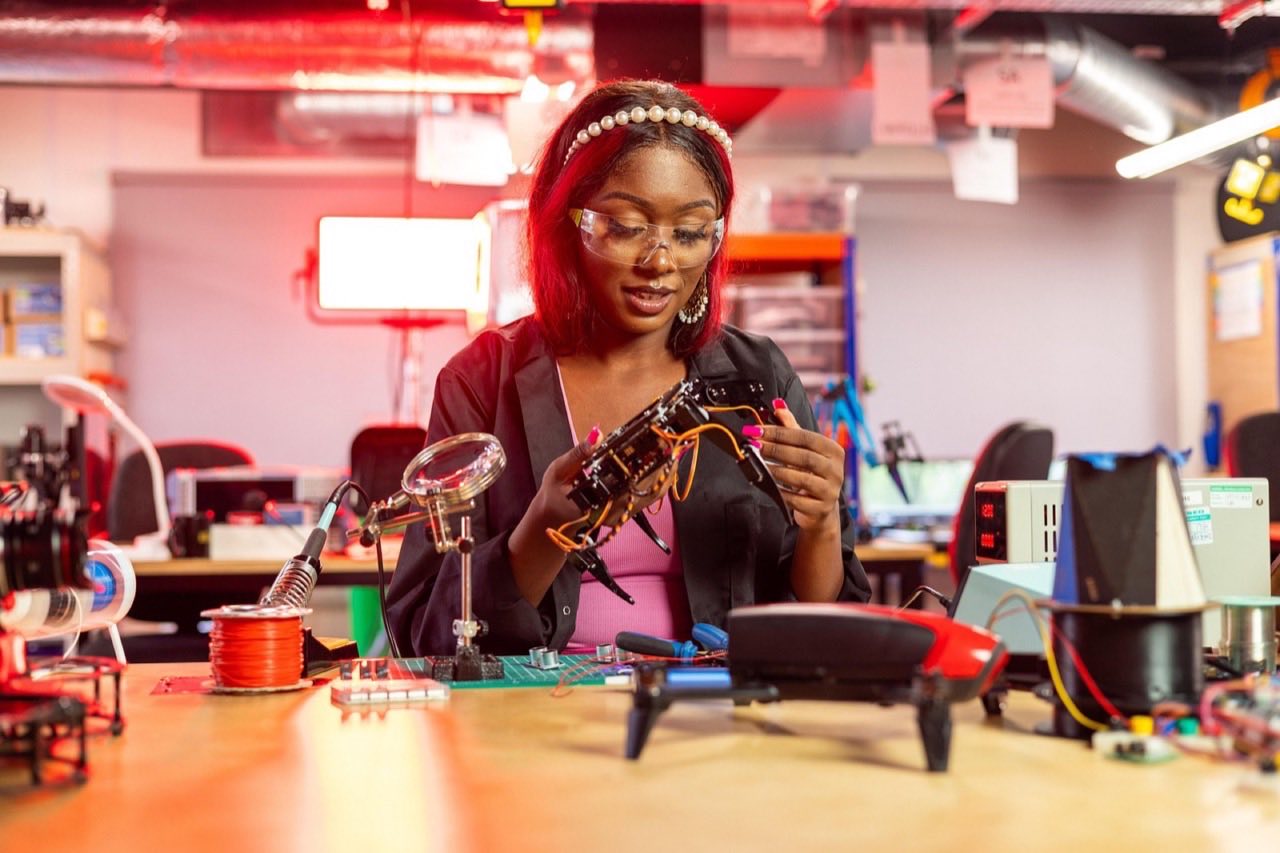 An engineer wearing safety glasses inspects a small electronics assembly on a lab bench, soldering tools nearby