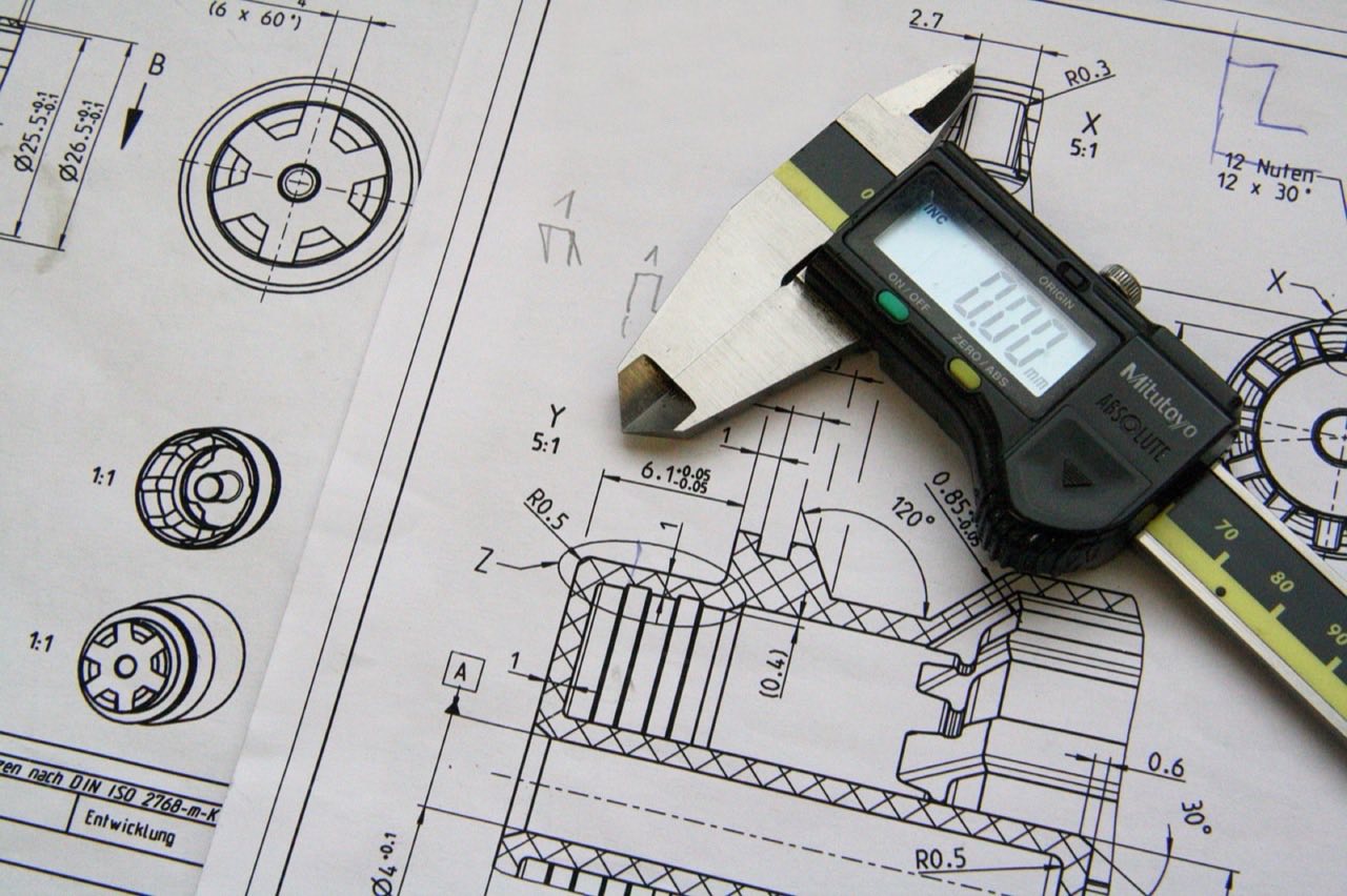 A digital caliper resting on a mechanical engineering technical drawing, set against dimensioned line work and section views