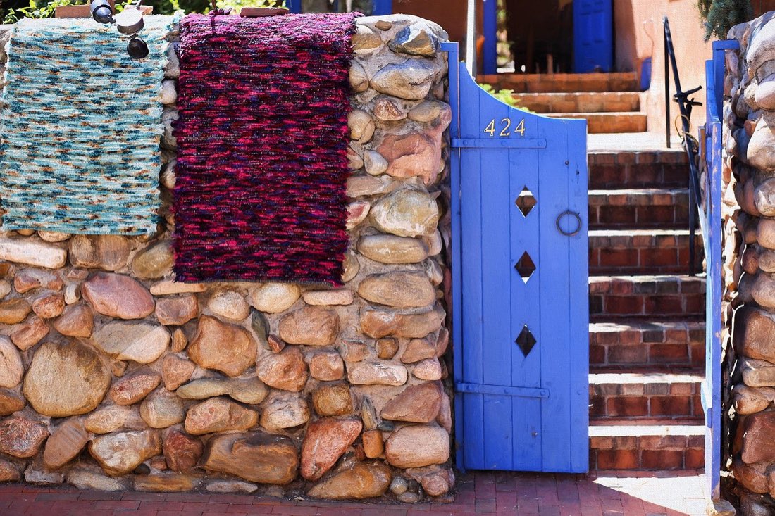 Hand-woven textiles hung to air over a stone courtyard wall beside a painted blue gate