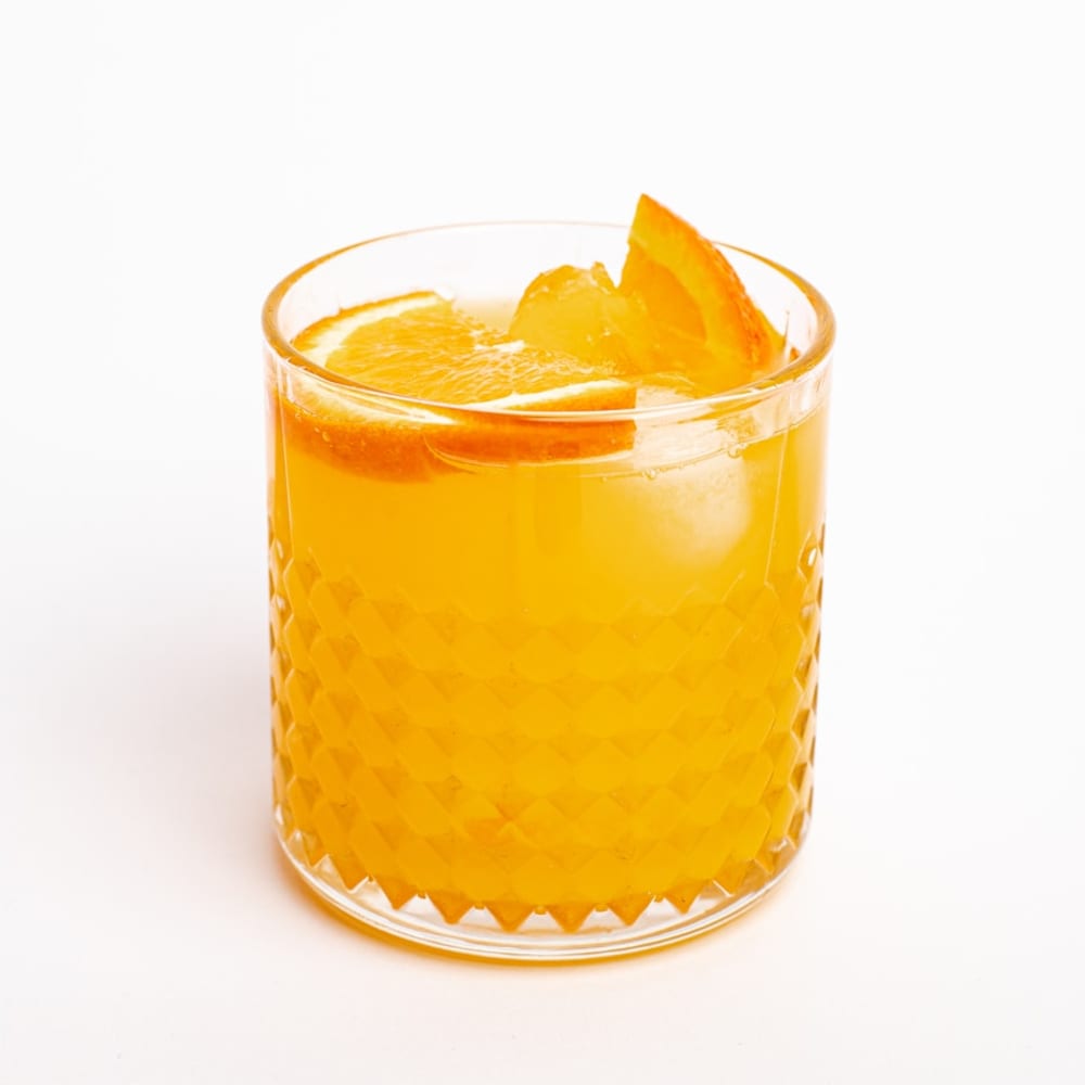 Hands holding a vibrant orange citrus surge juice against a white wall, glistening condensation on the glass