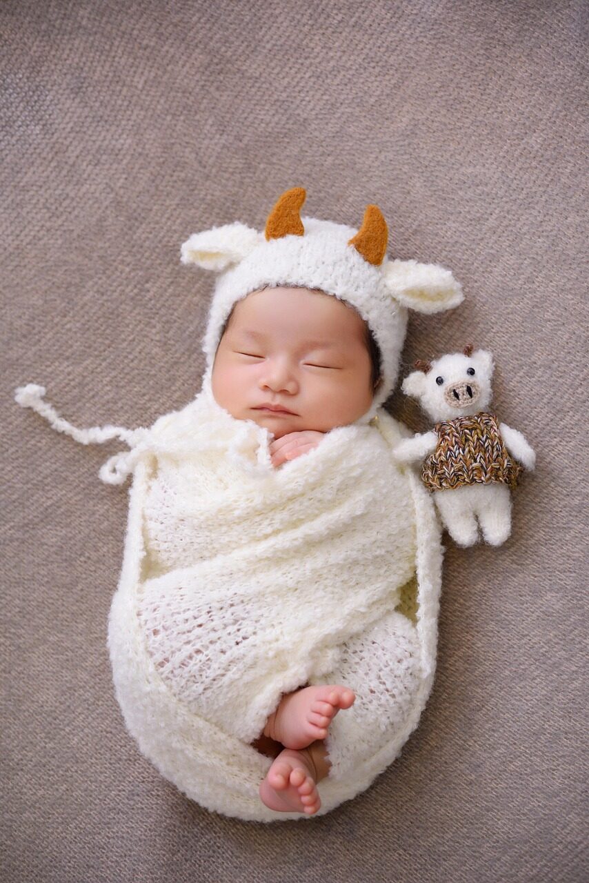 A sleeping newborn swaddled in cream knit wearing a bonnet with small ears, a knitted bear beside them