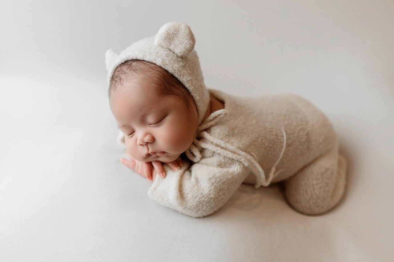 A sleeping newborn in a cream knitted romper and eared bonnet, curled with hands beneath the chin