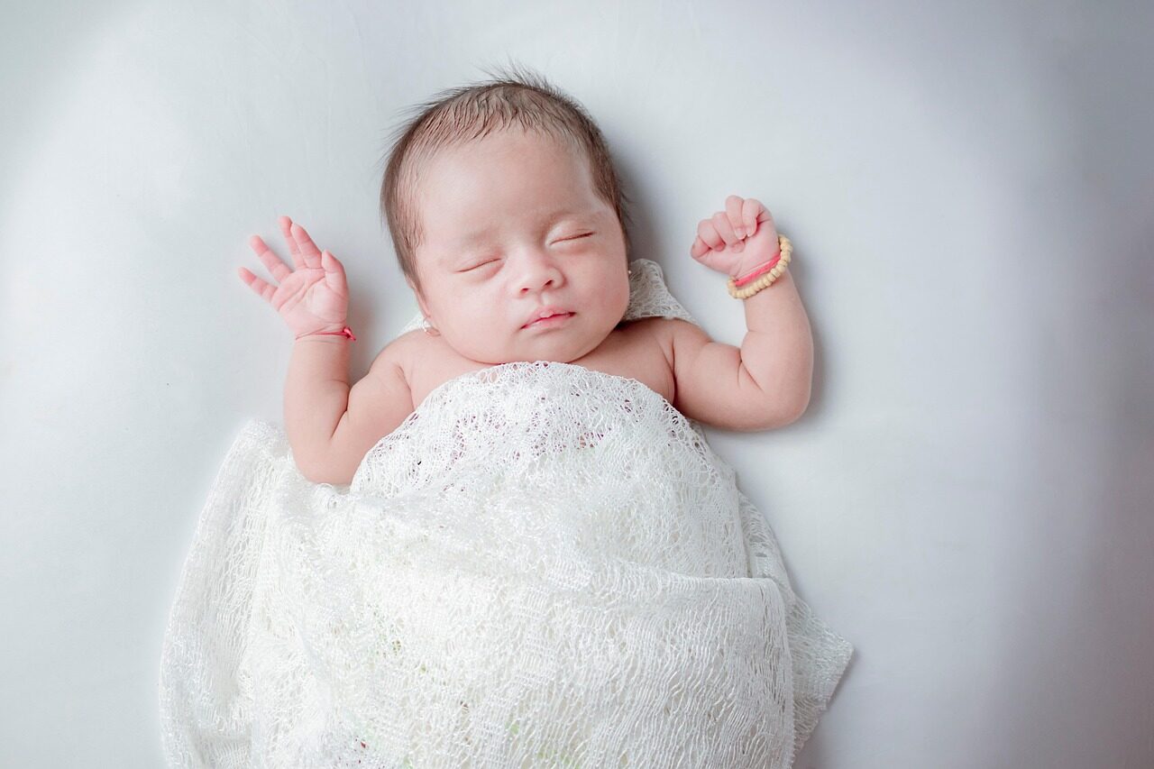 A sleeping newborn wrapped in white lace with both arms free, on a pale blue surface