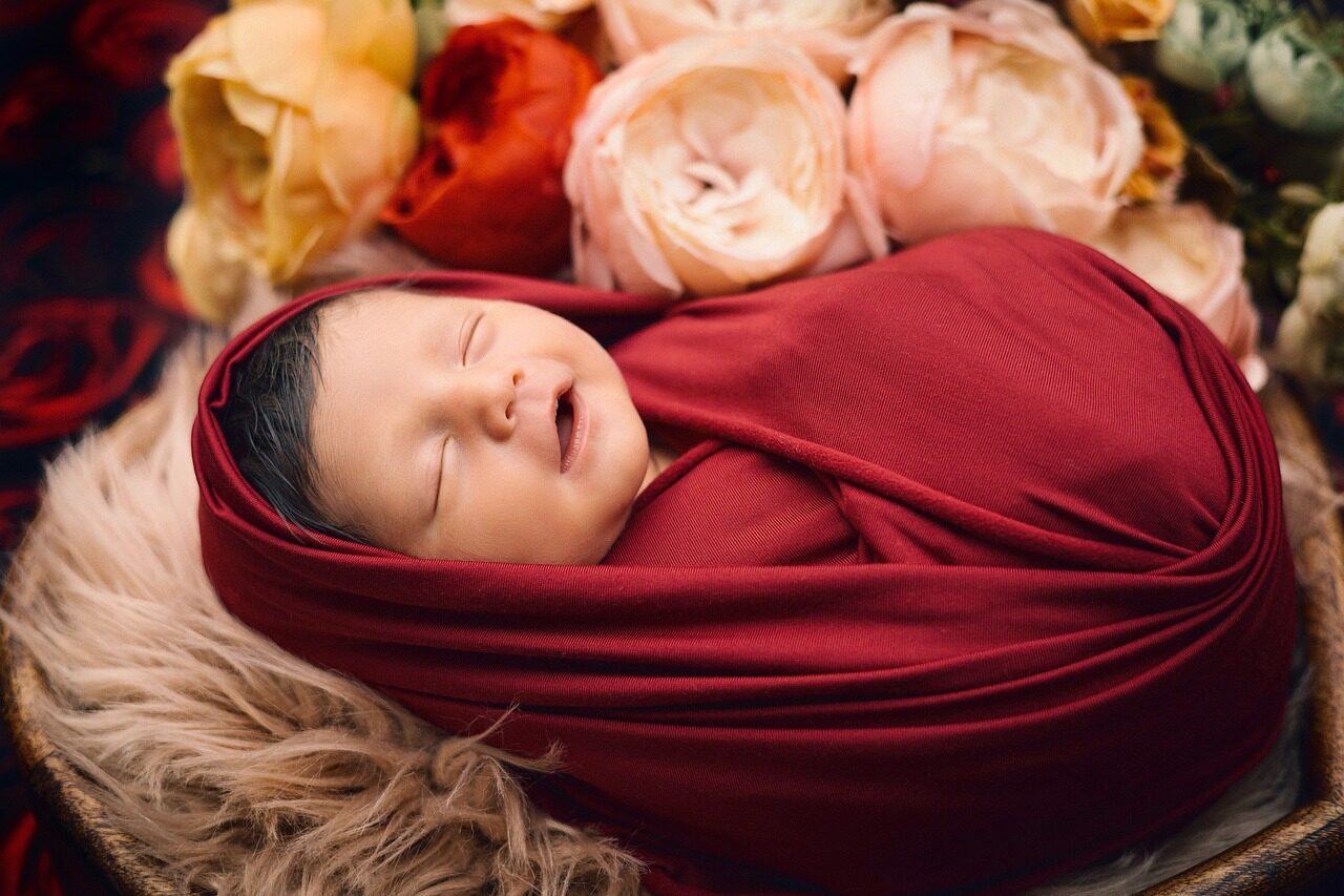A sleeping newborn swaddled in deep red cloth on cream fur, with pale roses behind