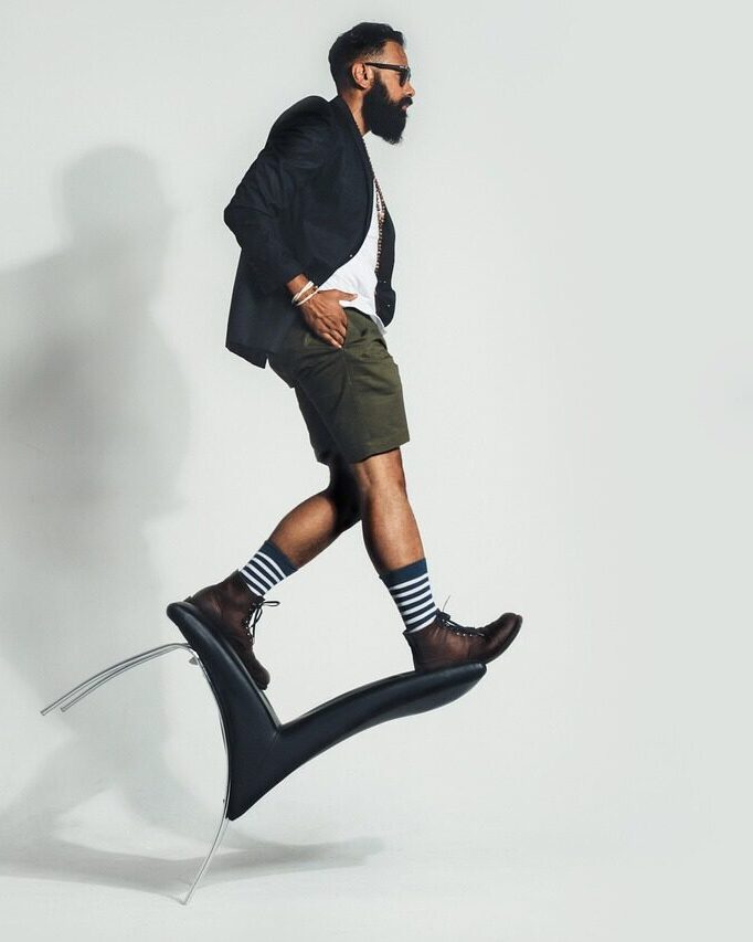 A bearded man in a blazer, shorts and boots balanced on a tipped chair, in profile against a white studio wall