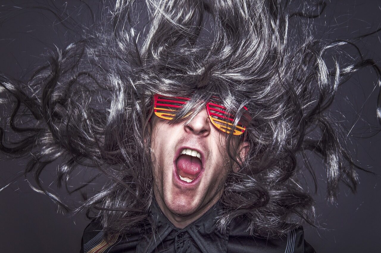 A person shouting with their long grey hair thrown outward, wearing slatted red and yellow novelty sunglasses