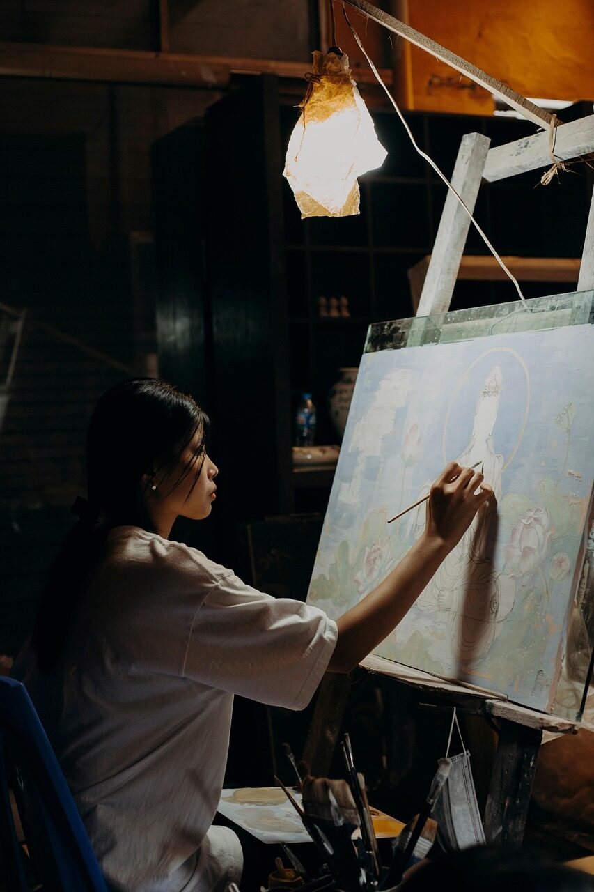A woman painting at an easel in a dim studio, seen from the side under a warm hanging lamp