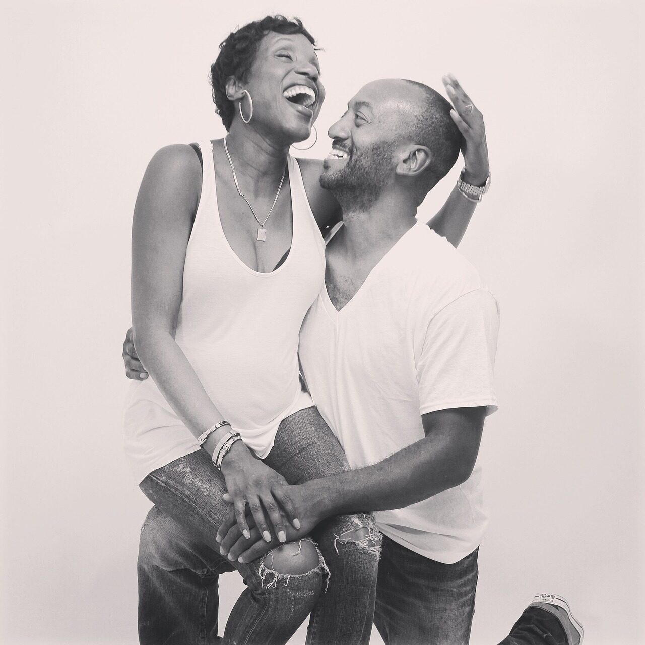 A couple in white vests and jeans laughing together in black and white, the man crouching beside the standing woman
