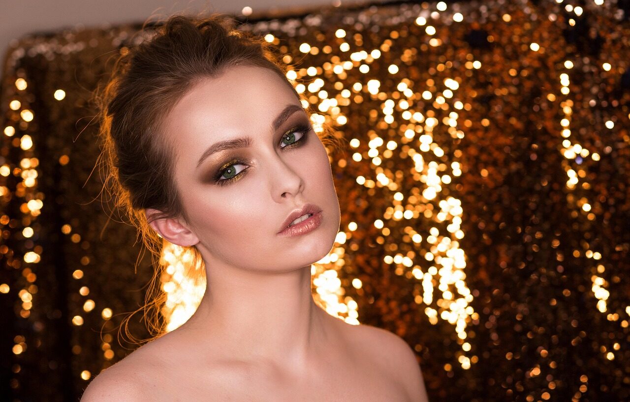 A woman with her hair up and strong eye make-up, lit against a background of gold glitter bokeh