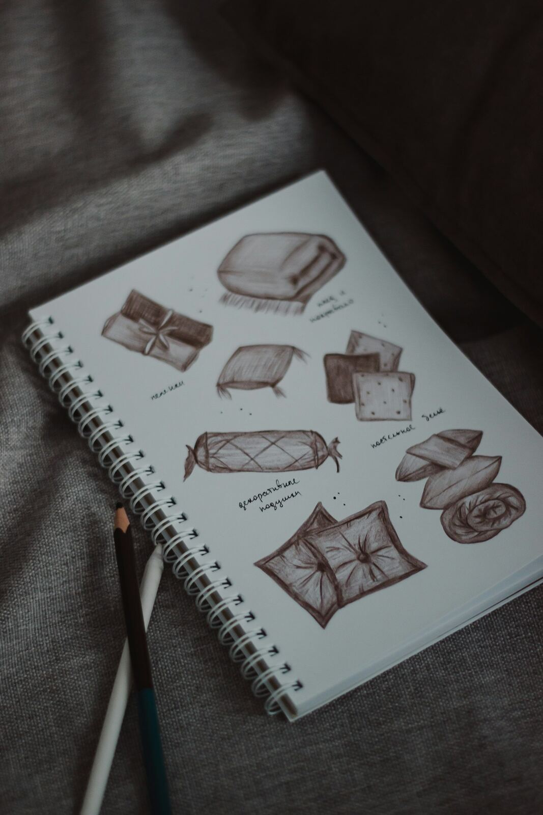 A spiral-bound sketchbook of pencil design drawings, resting on a grey fabric surface beside two pencils