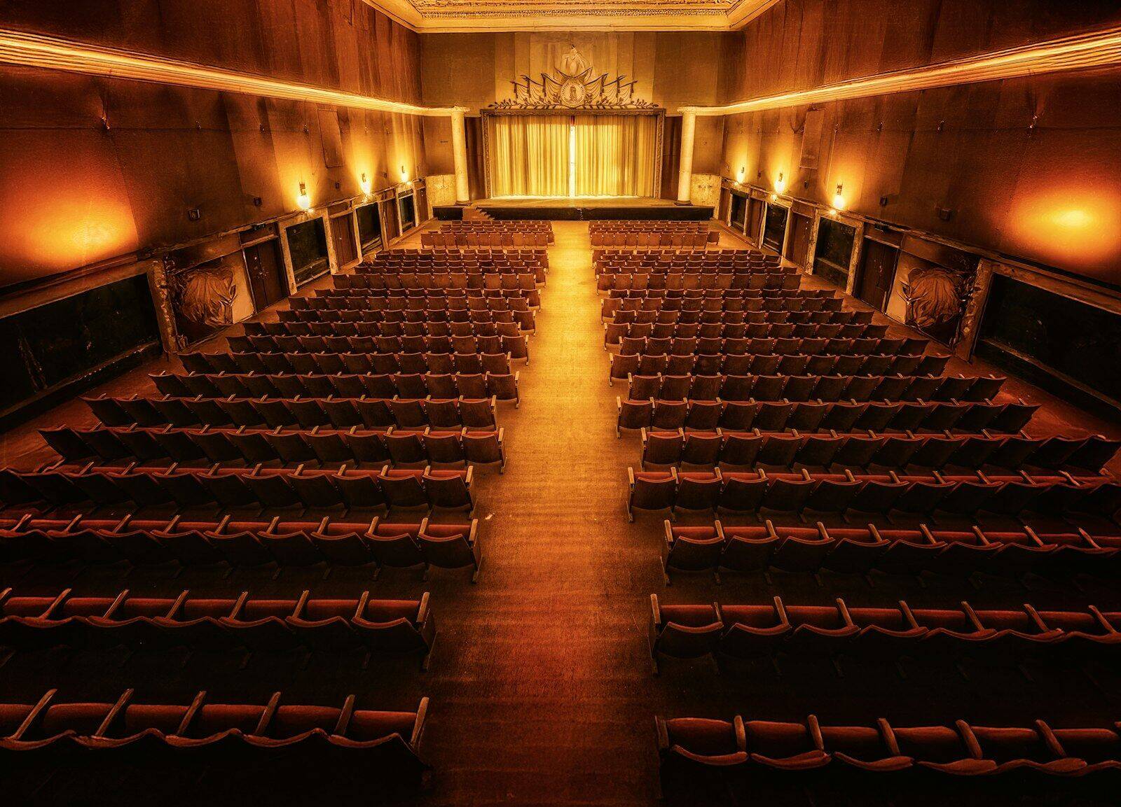 An empty, ornately decorated theatre auditorium with rows of red velvet seats facing a lit stage