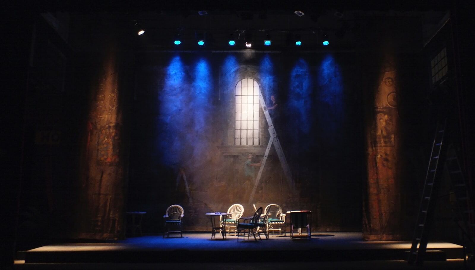 An empty stage set with a table and chairs beneath a tall painted window, lit in cold blue from above