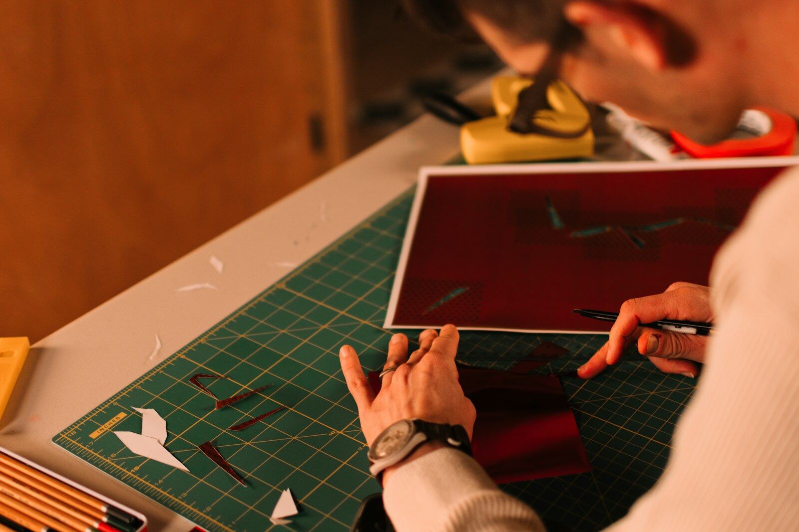 A pair of hands working on a paper craft project on a cutting mat, surrounded by pencils and tools