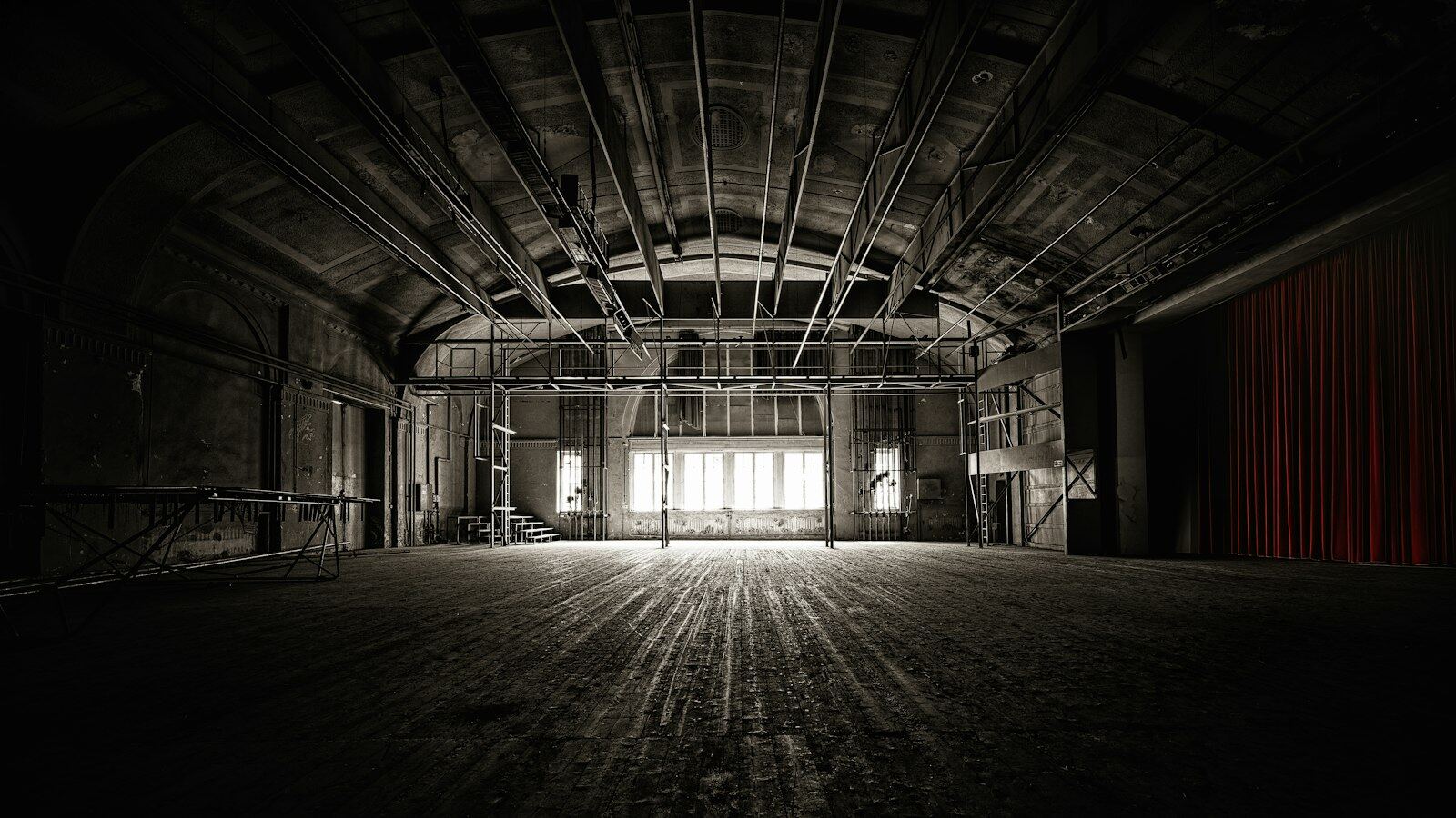 A dimly lit, cavernous empty theatre interior with exposed roof trusses, a rigging grid and a red curtain at the far end