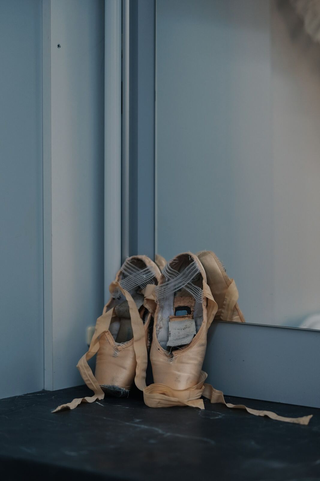A worn pair of pale pink ballet pointe shoes with ribbons, resting against a mirrored wall