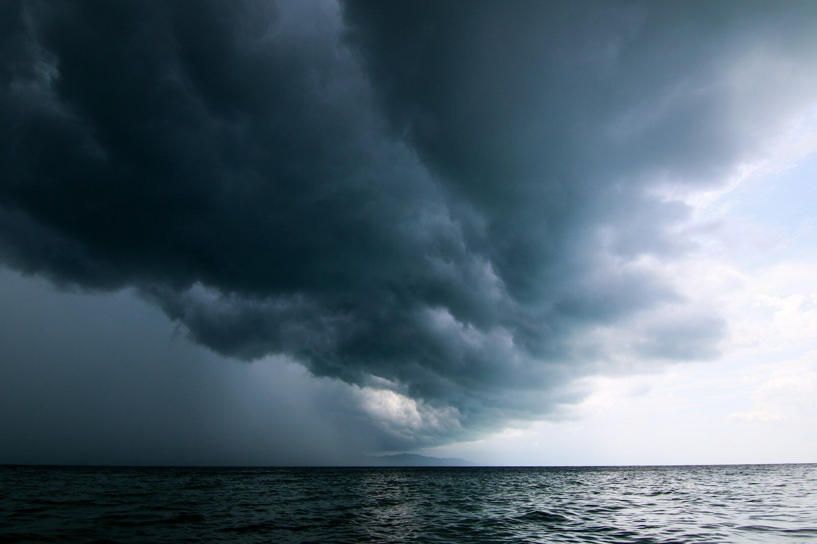 A large, dark storm cloud moving across the sky over open water, with a break of pale light at the horizon