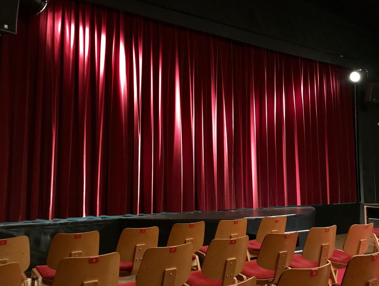 An empty auditorium: rows of wooden seats with red upholstery facing a lit red stage curtain.