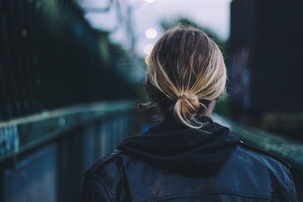 A woman photographed from behind in a black leather jacket crossing an urban footbridge