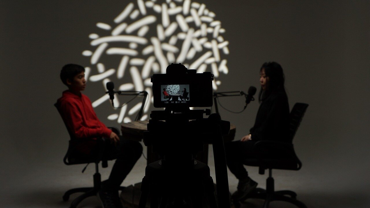 A studio interview two-shot: one person seated at the left edge of frame in a red hoodie, another at the right edge, with a camera on a tripod silhouetted between them against a patterned light projection.