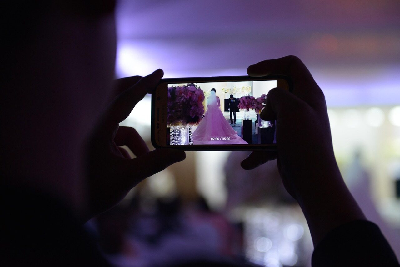 Hands holding a phone up to record, the bride blurred beyond it